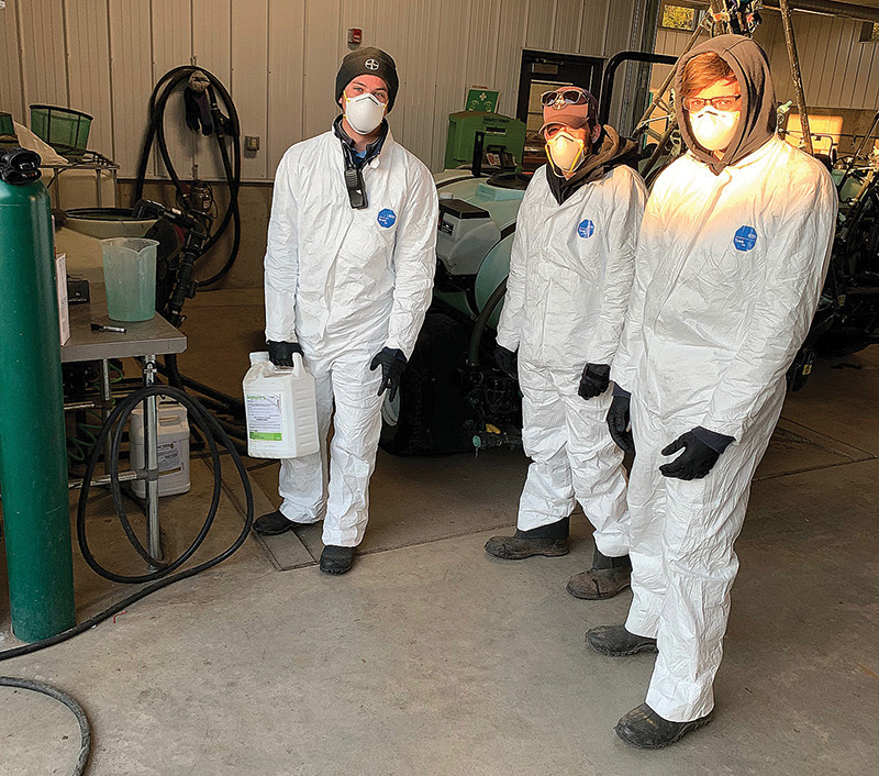 Three teenage boys wearing white hazmat suits as they work with chemicals in a maintenance shop at Lake Geneva Country Club.