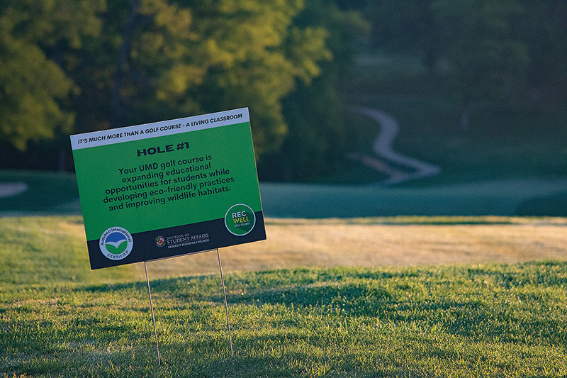 White, green and blue yard sign on a golf course. The sign reads: HOLE #1: Your UMD golf course is expanding education opportunities for students while developing eco-friendly practices and improving wildlife habitats. The bottom of the sign includes the logo for Audubon International.