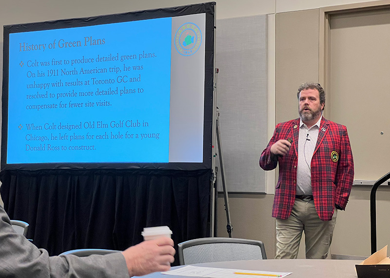 ASGCA member Nathan Crace stands in front of a group presenting on golf course design wearing the ASGCA's red plaid blazer uniform.