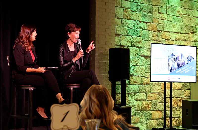 Two women sitting on a stage. The woman on the right is speaking into a handheld microphone