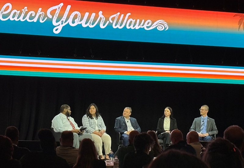 Four diverse men and women sit together on a stage in a hotel ballroom