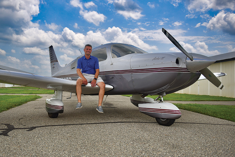 Scott Rettmann wearing a blue shirt, posing with a small gray airplane, which is parked outside a hangar