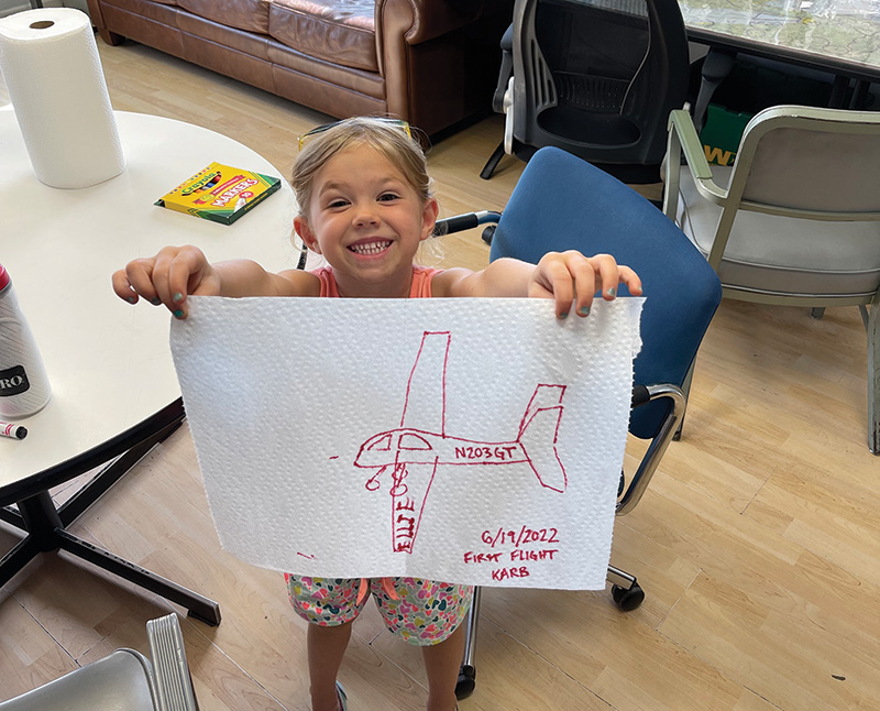 Ellie Rettmann, a young child in a floral dress, holds up a picture she has drawn in marker on a paper towel of her father in an airplane.