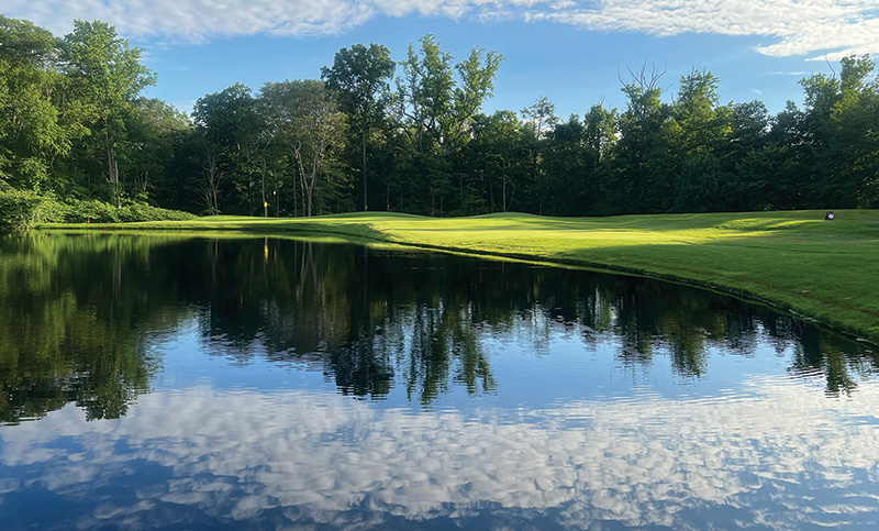 View of a clear golf course pond surrounded by a course and trees with the pond reflecting the sky