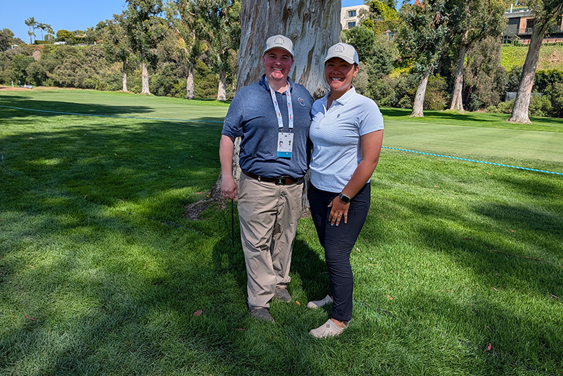 Two women wearing baseball caps stand in front of a eucalyptus tree on a sunny golf course