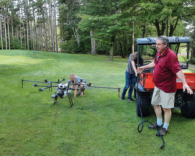 View of a golf course crew using a drone. A woman operates the drone in the background. A man stands in the foreground making space for it to take off and land.